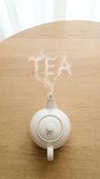 White ceramic teapot on wooden table with steam forming the word TEA, minimal Scandinavian style, warm cozy atmosphere for lifestyle and beverage concepts.