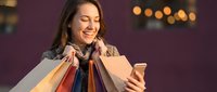 Happy young woman holding shopping bags while using smartphone with warm bokeh lights background for retail and consumer lifestyle concepts.
