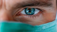 Close-up of medical worker's intense blue eye above surgical mask, showing determination and focus during healthcare procedure. Dramatic lighting highlights iris detail.
