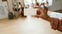 Hand holding blank white card at wooden desk with art supplies and sketches in background, suitable for business branding, stationery mockups, and creative workspace concepts.