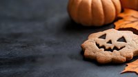 Halloween pumpkin-shaped gingerbread cookies with jack-o-lantern faces on dark surface with orange pumpkins and autumn leaves for seasonal baking and festive treats.