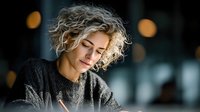 Young blonde curly-haired woman in gray sweater smiling peacefully with eyes closed in warm indoor lighting for lifestyle and wellness content.