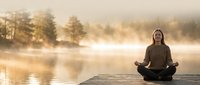 Young woman practicing yoga meditation in lotus pose on wooden dock at misty lake during golden sunrise creating peaceful wellness atmosphere.