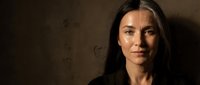 Professional portrait of confident young Caucasian businesswoman with long dark hair against textured brown wall background for corporate branding.