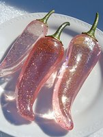 Three glossy pink chili peppers with green stems covered in sparkling glitter on white plate, artistic food styling for culinary design projects.