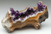 Raw amethyst crystal geode formation with deep purple crystals and natural brown agate bands, displaying natural mineral growth patterns on gray stone base.