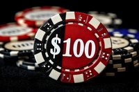 Casino poker chips stacked on dark background with hundred dollar denomination chip in focus, featuring red, black and white colors in dramatic lighting.