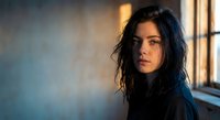 Young brunette woman with tousled hair in moody indoor lighting near window. Contemplative female portrait with dramatic shadows and warm golden light.
