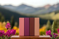 Books arranged in pink and burgundy shades standing on wooden surface against mountain landscape with vibrant wildflowers in soft evening light.