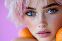 Close up portrait of young woman with pink hair, bright blue eyes and freckles holding oranges against purple background. Natural beauty makeup and fresh skin.
