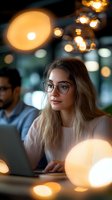 Young blonde businesswoman wearing glasses working on laptop in modern office with warm bokeh lighting and colleagues in background.