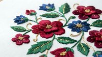 Colorful embroidered floral pattern with burgundy and blue flowers on white fabric. Traditional needlework craft featuring decorative blooms and green leaves.