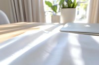 Soft morning sunlight casting shadows across white bedding, with laptop and houseplant creating minimal, peaceful home office atmosphere. Natural window lighting.
