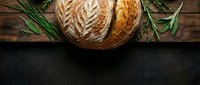 Artisan sourdough bread loaf with decorative wheat pattern scoring on rustic wooden background with fresh herbs for bakery and culinary designs.