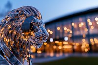 Glass lion sculpture with glowing city lights and bridge in background creates dramatic evening ambiance, highlighting transparent material and artistic craftsmanship.