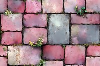 Pink and gray cobblestone pavement texture with small green plants growing between stones, creating rustic garden pathway pattern for landscape design.