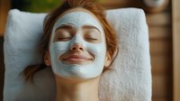 Young woman with red hair relaxing during spa facial treatment with clay mask, eyes closed and gentle smile, lying on white towel in wellness center.