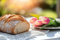 Fresh crusty artisan bread loaf on white lace doily with pink spring tulips in soft natural lighting, creating romantic breakfast or bakery atmosphere.