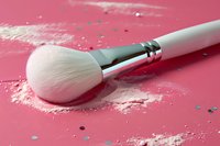 White professional makeup brush with scattered loose face powder on pink background, close-up view showing cosmetic application tools and beauty products.