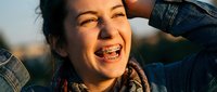 Young woman with dental braces smiling joyfully outdoors in golden sunlight, hands behind head showing confidence and happiness during orthodontic treatment.