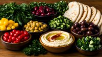Hummus with fresh vegetables, olives, pita bread and herbs on wooden table. Mediterranean appetizer platter with colorful ingredients for healthy eating.