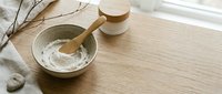Flat lay composition with natural skincare ingredients, wooden spoon in ceramic bowl with white powder, cream jar, and dried botanicals on light wood surface for cosmetic and wellness branding.