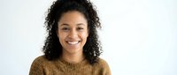Young African American woman with curly hair smiling warmly in brown knit sweater against white background for lifestyle and portrait photography.