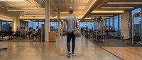 Young athletic man walking through modern fitness center with floor-to-ceiling windows and professional gym equipment for workout motivation and healthy lifestyle.