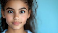 Young mixed race girl with curly dark hair and bright blue eyes smiling confidently against turquoise background, natural beauty portrait showing positive emotion.