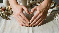 Female hands forming heart shape over natural linen fabric with dried flowers, expressing love and care in rustic home setting for wellness concepts.