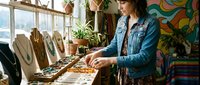 Female artisan with dark hair and medium complexion working at wooden jewelry display table in bohemian studio, arranging handmade necklaces and accessories. Denim jacket, natural daylight from windows, colorful wall art, potted plants, and textile backdrop create creative workspace atmosphere for small business, craft entrepreneurship, and artisan jewelry making.