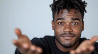 Young African American man with curly hair and friendly smile gesturing with open palms against gray background, expressing welcoming and positive attitude.