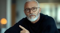 Confident mature businessman with gray beard and glasses wearing black sweater gestures thoughtfully during professional conversation in modern office setting.