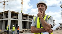 Thoughtful senior construction manager in safety vest and hard hat overseeing building site with cranes and workers in background during development project.