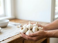 Fresh garlic bulbs held in hands over wooden kitchen counter with white bowl and natural window light, suitable for cooking, recipe, and food preparation content.
