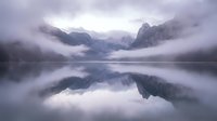 Misty mountain lake with perfect reflections in calm water surrounded by dramatic peaks shrouded in fog creating serene atmospheric landscape scene.