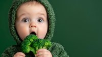 Surprised baby in green knitted hoodie eating fresh raw broccoli floret against dark green background. Healthy eating habits concept for toddlers.