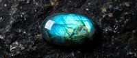 Polished labradorite gemstone with brilliant blue-green flash on dark textured surface. Natural mineral specimen displaying iridescent schiller effect for jewelry making.