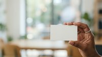 Person with dark skin tone holding blank white business card in modern office environment with blurred background, suitable for corporate branding and professional marketing materials.