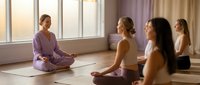Female yoga instructor leading meditation class with diverse women students sitting cross-legged on mats in bright studio with large windows.
