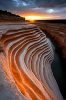Dramatic sunset illuminates layered sandstone formations with warm orange light, creating striking patterns and textures along canyon walls against stormy sky.