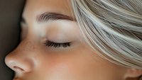 Close up view of blonde woman's eye area showing natural makeup, long eyelashes, and well-groomed eyebrow against soft blurred background.