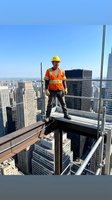 Construction worker in yellow hard hat and orange safety vest standing on high-rise building beam above cityscape. Use for safety, construction, and workplace themes.