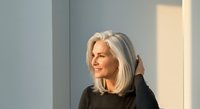 Senior woman with long silver hair wearing black sweater, standing by window with natural light, mature professional portrait for business, healthcare, lifestyle and corporate use.