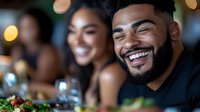 Young African American man with beard laughing heartily at restaurant gathering with friends, genuine joy and happiness during social dinner party celebration.