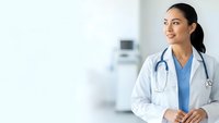 Young female doctor in white coat with stethoscope standing confidently in modern hospital setting with medical equipment background.