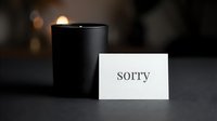 Small white card with the word sorry leaning against a dark candle on a moody gray surface, conveying apology, sympathy, or condolence.