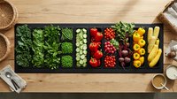 Assorted fresh vegetables arranged in black compartments on wooden surface, including leafy greens, tomatoes, peppers, and beans for healthy cooking and meal planning.
