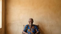 Senior African man wearing blue patterned traditional shirt and eyeglasses, seated against warm beige textured wall, suitable for cultural portraits and diversity stock imagery.