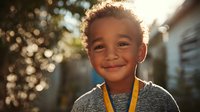 Young African American boy with curly hair smiling warmly at camera wearing gray shirt with yellow lanyard in outdoor setting with soft bokeh lighting.